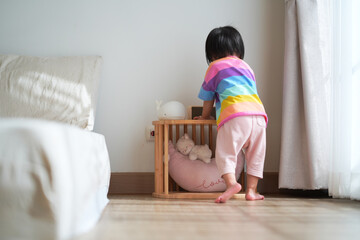 A young child wearing a colorful striped shirt kneels on the wooden floor in a cozy bedroom, looking toward a small wooden crib with soft toys.
