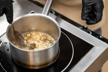 A professional chef stirs risotto or rice soup with a spoon in a stainless saucepan on an induction cooktop; focus on food preparation cooking