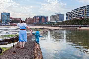 mum with young Aussie kid in swimmers walking beside the canoe pool on the coast at Newcastle Beach