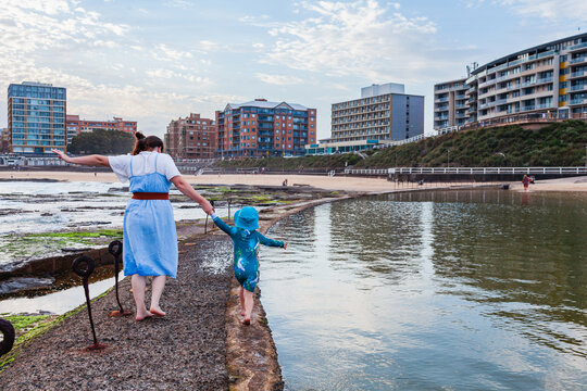 mum with young Aussie kid in swimmers walking beside the canoe pool on the coast at Newcastle Beach