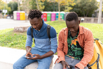 Young african friends connecting with mobile devices while sitting on a low wall outdoors
