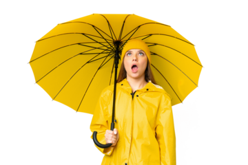 Young blonde woman with rainproof coat and umbrella over isolated chroma key background looking up and with surprised expression