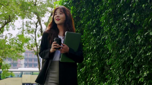 Young businesswoman walking outdoor, traveling with laptop, near modern office building, enjoying daily commute with beverage, amidst green urban landscape, projecting success, work life balance.