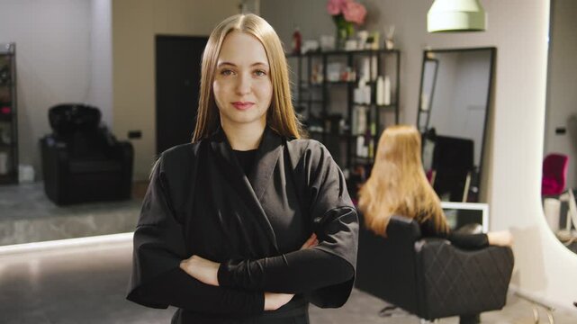 Professional hair stylist in salon with sleek hair, wearing black outfit, confidently posing in modern workspace with client in background