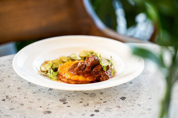 A close-up shot of slow-cooked beef stew served over a golden polenta cake, garnished with fresh microgreens and sliced pickles on a white plate in a restaurant setting
