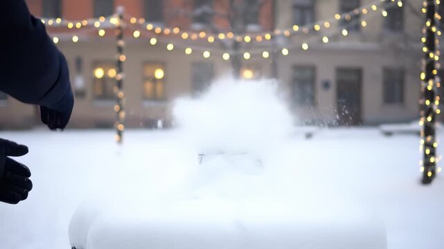 Hands in navy gloves prepare to throw a snowball in a winter wonderland, with twinkling lights illuminating the snowy scene, capturing the joy of seasonal playfulness