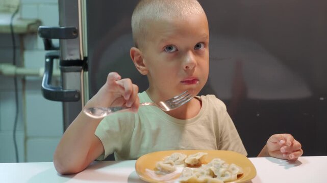 Hot meat dumplings with sour cream fill the yellow plate on the table. The hungry child is holding a silver fork to eat the savory pastry, sticking his tongue out in anticipation.