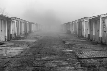 Zamglona aleja starych garaży w czerni i bieli. Monochromatyczna fotografia przedstawiająca perspektywę rzędu starych, betonowych garaży spowitych gęstą mgłą. © Henryk Niestrój