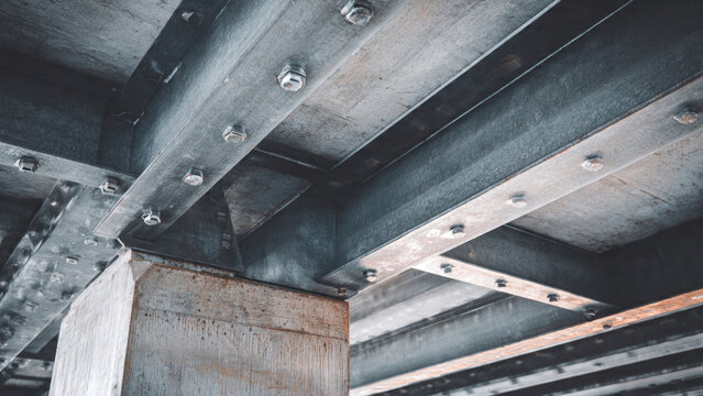 Close-up of an industrial structure featuring robust steel beams, cross-members, and a concrete support column, all secured with numerous bolts from below.