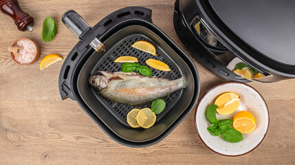 Whole fresh fish with lemon slices and basil prepared in an air fryer basket on a wooden table, top view, copy space