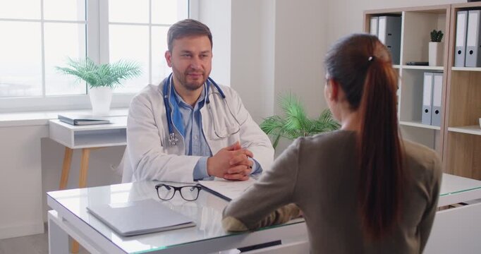 Young friendly doctor consulting woman patient sitting at desk on workplace in examination room in medical clinic and giving recommendations in treatment in office. 4k video. slow motion video.