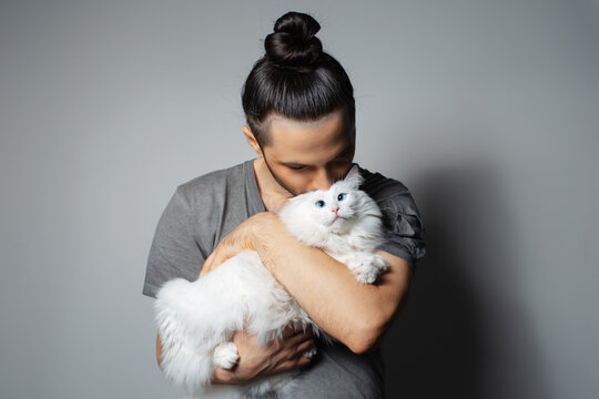 Studio portrait of young man with white cat in arms while kissing the pet on grey background.