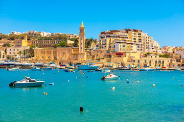 Fishing village and resort town of Marsaskala, Malta, nestled around turquoise bay. Numerous boats bobbing in harbor water framed by hillside buildings and notable bell tower of St. Anne Parish Church
