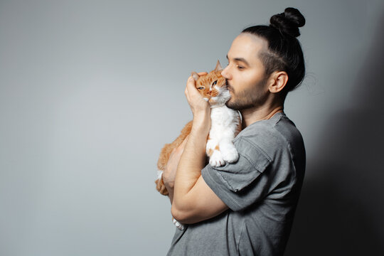 Studio portrait of young man with red cat in arms while kissing the pet on grey background.