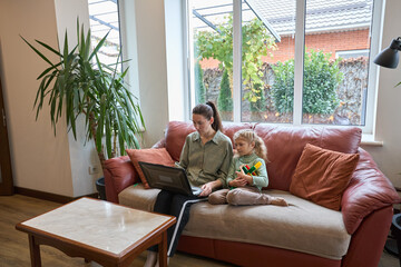 Mother and daughter relax on cozy sofa with laptop in bright living room