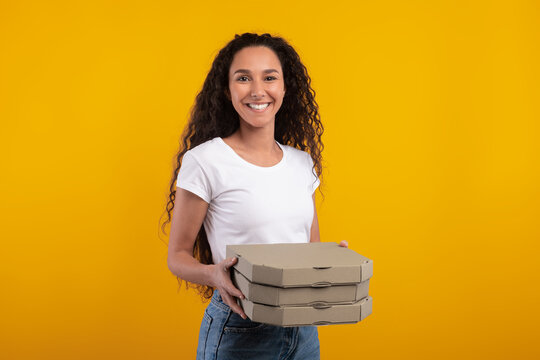 Excited young woman stands against a yellow orange background holding a stack of pizza boxes. She beams with joy, highlighting the fun of enjoying a cheat meal or fast food delivery. - Powered by Adobe
