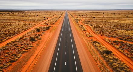 High angle aerial view of a long, straight asphalt highway stretching to the horizon through a vast, remote outback landscape with red earth and sparse vegetation under an overcast sky