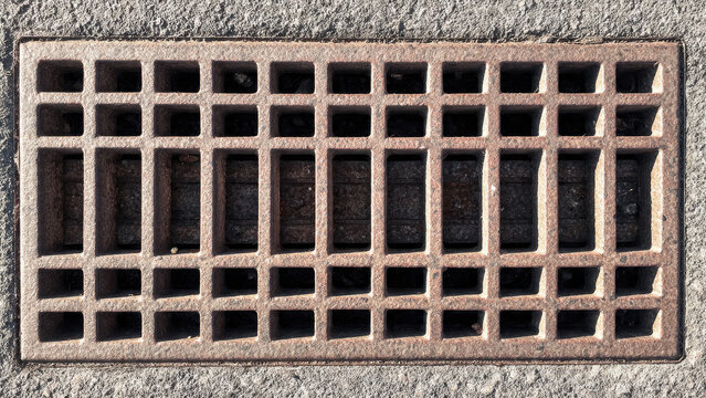 A weathered, rusty rectangular metal grate with a grid pattern, likely for drainage, set into rough concrete pavement.
