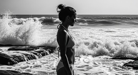 Contemplative woman in a bikini standing in profile on a rocky shoreline, looking at the powerful ocean waves in a dramatic black and white artistic photograph