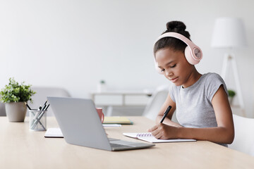 A young black girl is studying intently in her living room. She is wearing headphones and taking notes while using her laptop. This scene reflects remote learning during the pandemic.
