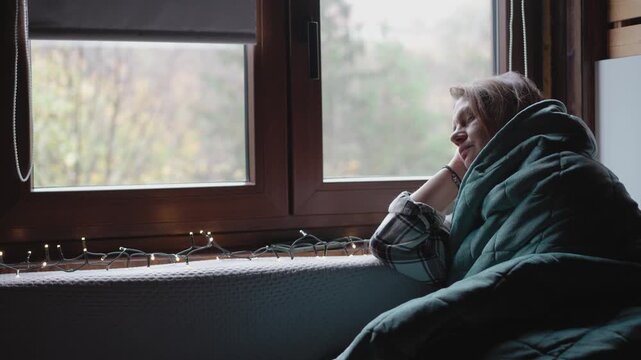 An adult woman warms up by wrapping herself in a warm blanket while sitting by the window of a country house