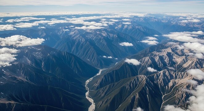 A majestic aerial view of a vast mountain range with snow-capped peaks and a winding river valley seen through scattered clouds on a sunny day