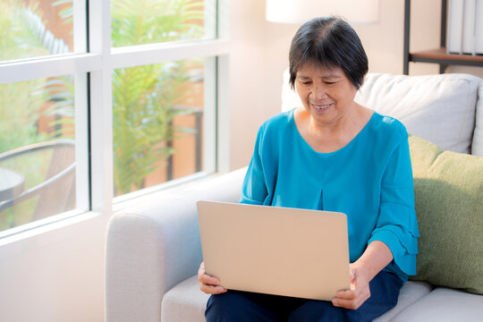 Asian senior woman sitting on sofa using laptop computer for looking social media in living room at home, elderly doing activities and communication, relaxation and leisure of retirement.
