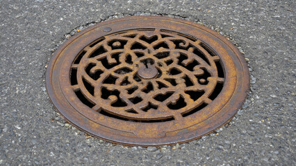 A weathered, circular, rusty manhole cover with an intricate decorative pattern is set within dark grey asphalt pavement, featuring a central keyhole. It's an aged urban utility fixture.