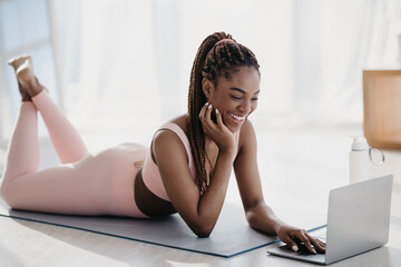 A joyful young African American woman lies on a mat at home, engaging with her personal coach on a laptop during an online fitness class. She enjoys following the workout.