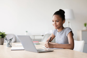 A cheerful teenage girl is studying at home during an online lesson. She takes notes while focusing on her laptop in a cozy living room. Learning from home makes her engaged and happy.