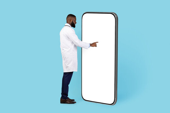 African American Doctor Standing Near Big Blank Smartphone And Touching White Screen, Young Black Male Physician Browsing New Mobile Application While Standing On Blue Background, Collage, Mockup