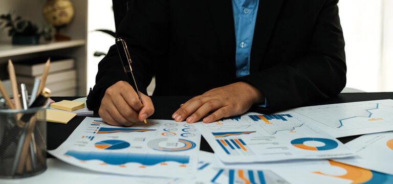 A business professional engaged in a detailed analysis of financial reports and graphs at a modern office desk, showcasing focus and commitment to data-driven decisions.Noogenesis