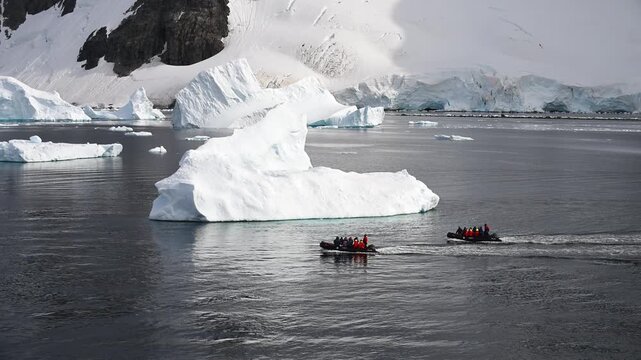 A group of adventurers takes an inflatable boat to explore towering icebergs in a stunning arctic landscape. The water is calm, reflecting the blue ice under a clear sky.