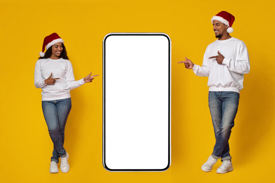 Two individuals in festive attire stand on either side of a large blank smartphone. They are smiling and pointing, celebrating the holiday spirit against a vibrant yellow backdrop. - Powered by Adobe