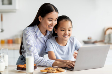 A happy Asian mother and her child sit at the kitchen table, engaging with a laptop. They enjoy homemade pastries while spending quality time together in a cozy home setting.