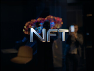A metallic three dimensional NFT text stands sharply in focus against a dark blurred background where colorful abstract lights glow and a person holding a smartphone appears out of focus