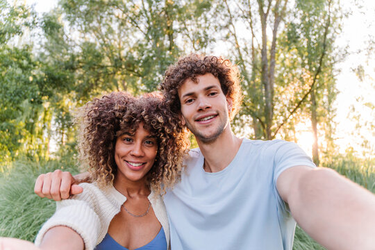 Happy young couple with curly hair smiling and looking at the camera, taking a selfie outdoors in a park during sunset, reflecting friendship, togetherness, and joy