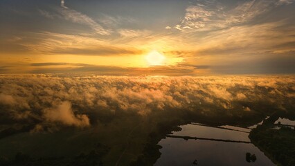 A high-altitude aerial photograph capturing a sunrise above a vast forest with golden morning mist drifting through the treetops