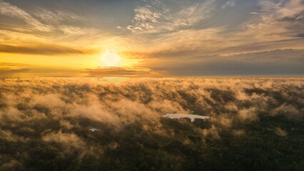 A high-altitude aerial photograph capturing a sunrise above a vast forest with golden morning mist drifting through the treetops