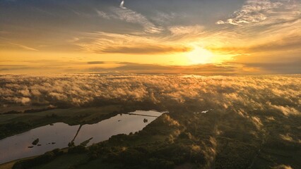A high-altitude aerial photograph capturing a sunrise above a vast forest with golden morning mist drifting through the treetops