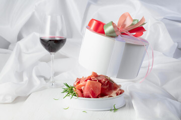 Prosciutto with rosemary in gift box and glass of red wine on a white wooden table.