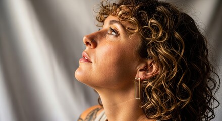 Serene profile portrait of a thoughtful young woman with curly hair, freckles, and a nose piercing looking upwards with a hopeful gaze against a neutral studio background