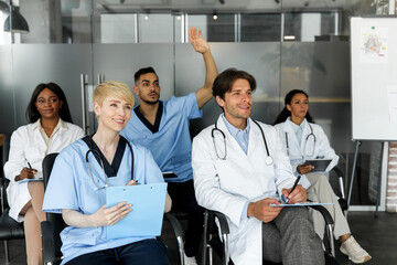 Multiracial team of doctors sitting at hall or clinic cabinet, listening to speech, having professional training or attending medical conference, young middle-eastern doctor raising hand up