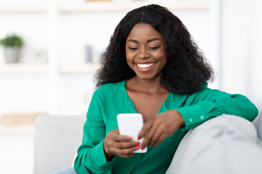 A young woman smiles as she interacts with her smartphone, sitting on a cozy couch in a well-lit living room. The atmosphere feels relaxed and cheerful.