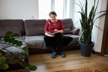 Elderly woman reading a book on sofa in cozy living room with plants