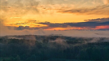 A high-altitude aerial photograph capturing a sunrise above a vast forest with golden morning mist drifting through the treetops