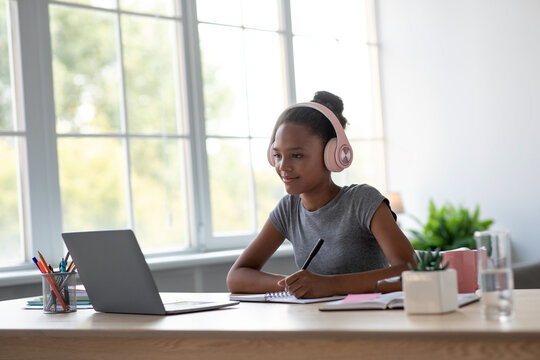 Glad teenage African American lady student focuses on her online lessons while studying at home. She is taking notes and using her laptop by a large window.
