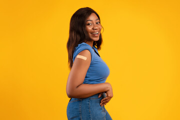 Obraz premium A black young woman stands in a studio with a bright yellow background, showing her vaccinated arm with a plaster after receiving a Covid-19 injection. Her expression is joyful and confident.
