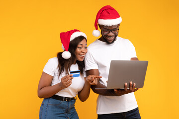 Couple celebrating the festive season while shopping online. They are focused on their laptop, using a credit card to buy gifts. The backdrop is a cheerful yellow in a studio setting.