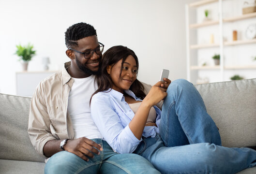 Young African American couple sits on a comfortable couch, smiling as they look at a cellphone. The man wears glasses and braces, and they share a joyful moment while exploring social media together.
